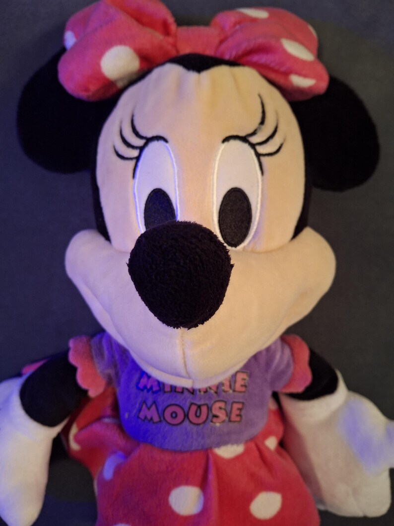 May include: A plush Minnie Mouse doll with a pink bow with white polka dots. The doll has black ears, a black nose, and large eyes. She wears a purple top with the words "MINNIE MOUSE" and a red dress with white polka dots.