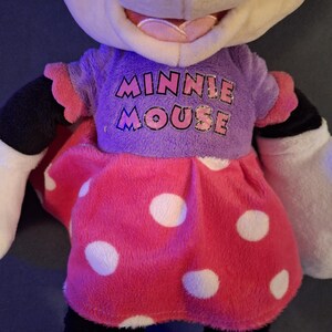May include: A plush Minnie Mouse doll. The doll has a purple shirt with the words "MINNIE MOUSE" in pink and blue. The doll is wearing a pink dress with white polka dots. The doll has black legs, white gloves, and black ears.