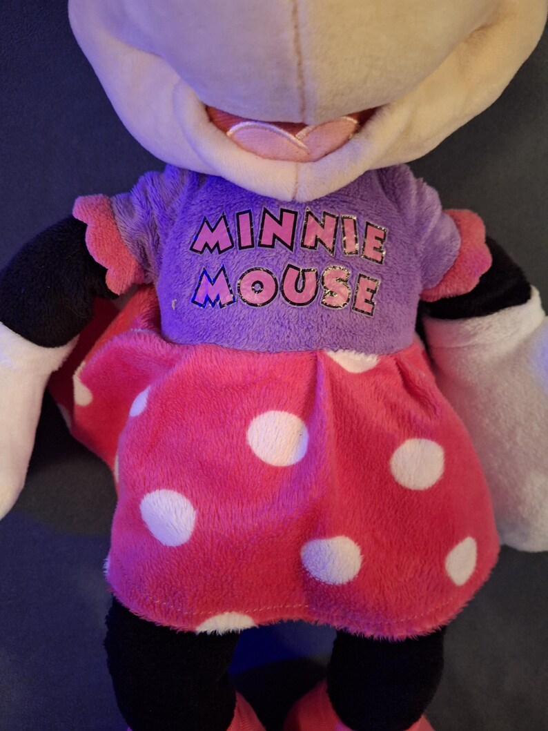 May include: A plush Minnie Mouse doll. The doll has a purple shirt with the words "MINNIE MOUSE" in pink and blue. The doll is wearing a pink dress with white polka dots. The doll has black legs, white gloves, and black ears.