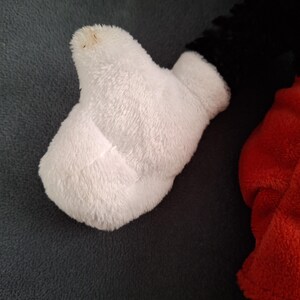 May include: Close-up of a white plush glove, part of a larger stuffed toy. The glove is made of soft, fluffy material. The black and red parts of the toy are visible in the background. The glove has a small brown spot on the tip.