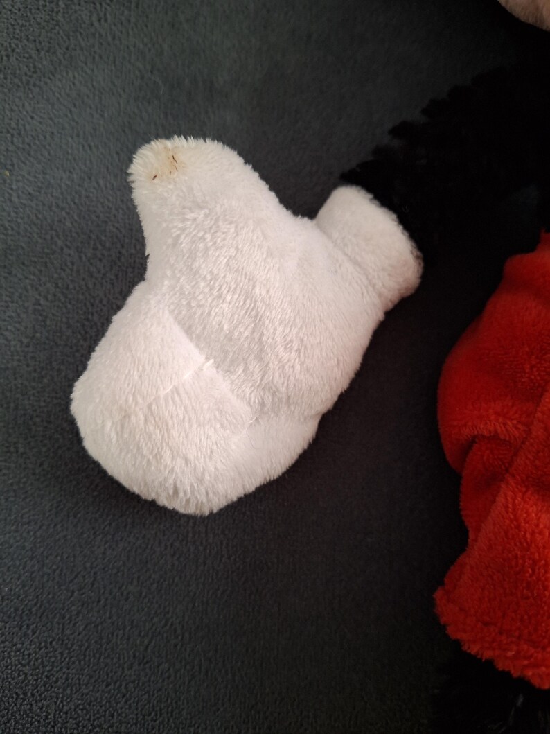 May include: Close-up of a white plush glove, part of a larger stuffed toy. The glove is made of soft, fluffy material. The black and red parts of the toy are visible in the background. The glove has a small brown spot on the tip.