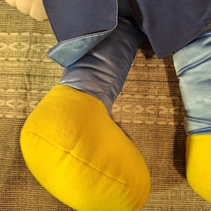 May include: Close-up of a plush toy's legs and lower body. The legs are a light blue satin material, with yellow feet. The toy's outfit includes a navy blue jacket.
