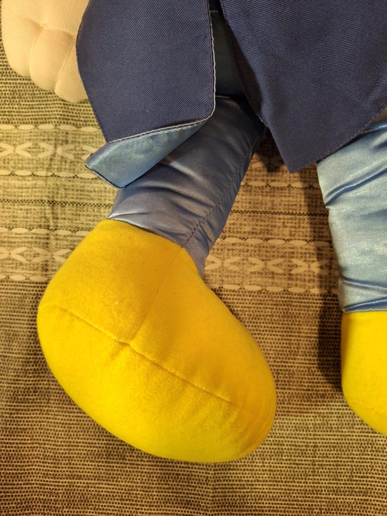 May include: Close-up of a plush toy's legs and lower body. The legs are a light blue satin material, with yellow feet. The toy's outfit includes a navy blue jacket.