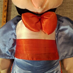 May include: A plush doll featuring a black and white head with a pink nose and a large orange bow tie. The doll is wearing a light blue satin outfit with an orange sash.