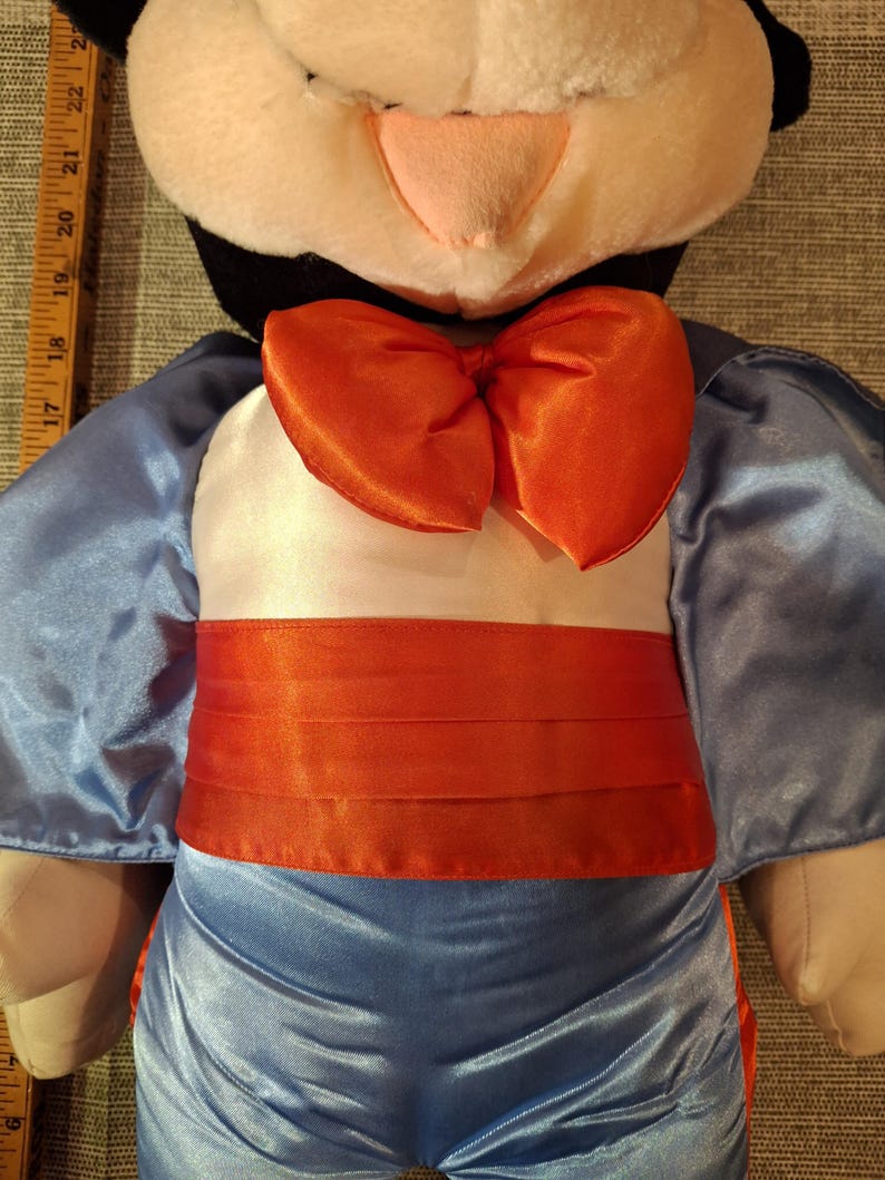 May include: A plush doll featuring a black and white head with a pink nose and a large orange bow tie. The doll is wearing a light blue satin outfit with an orange sash.