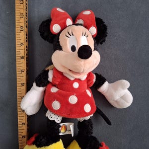 May include: A plush Minnie Mouse doll, featuring a red dress with white polka dots, a matching bow, and yellow shoes. The doll has black fur, white gloves, and a smiling face. The doll is approximately 12 inches tall.