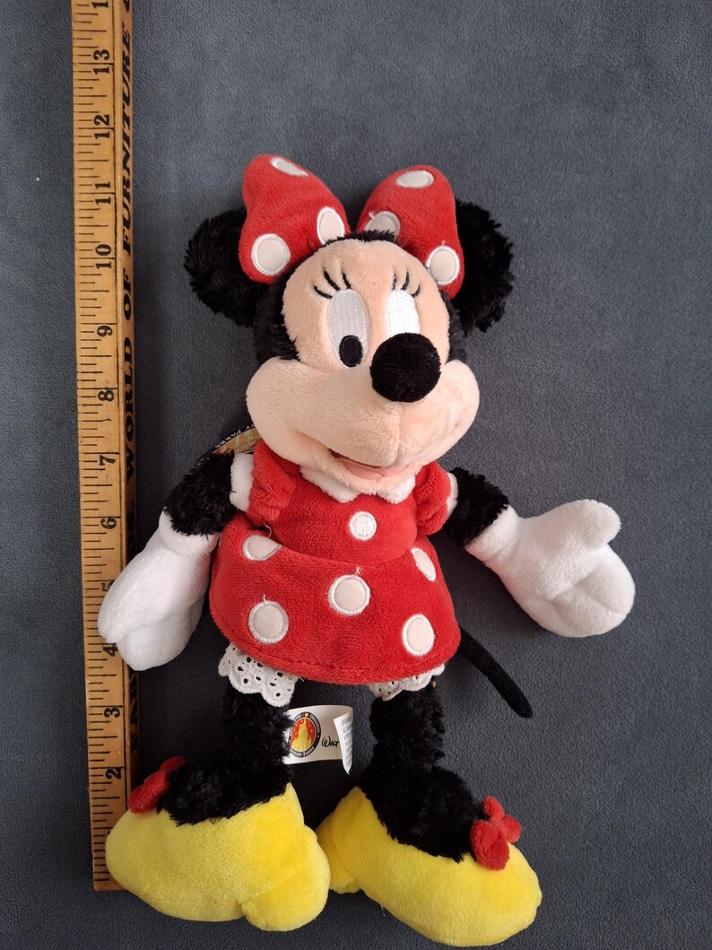 May include: A plush Minnie Mouse doll, featuring a red dress with white polka dots, a matching bow, and yellow shoes. The doll has black fur, white gloves, and a smiling face. The doll is approximately 12 inches tall.