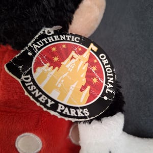 May include: A plush Mickey Mouse toy with a red shirt, black fur, and white gloves. The tag reads "Authentic Disney Parks Original" with a castle graphic. The toy's features include a black head and ears.