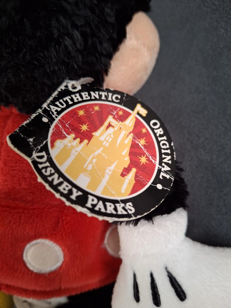 May include: A plush Mickey Mouse toy with a red shirt, black fur, and white gloves. The tag reads "Authentic Disney Parks Original" with a castle graphic. The toy's features include a black head and ears.