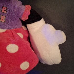 May include: A plush doll featuring a red polka-dot skirt, a purple top with the letter "E" on it, and a white glove. The doll's arm is black and white, with a pink ruffled sleeve. The doll is a popular character.