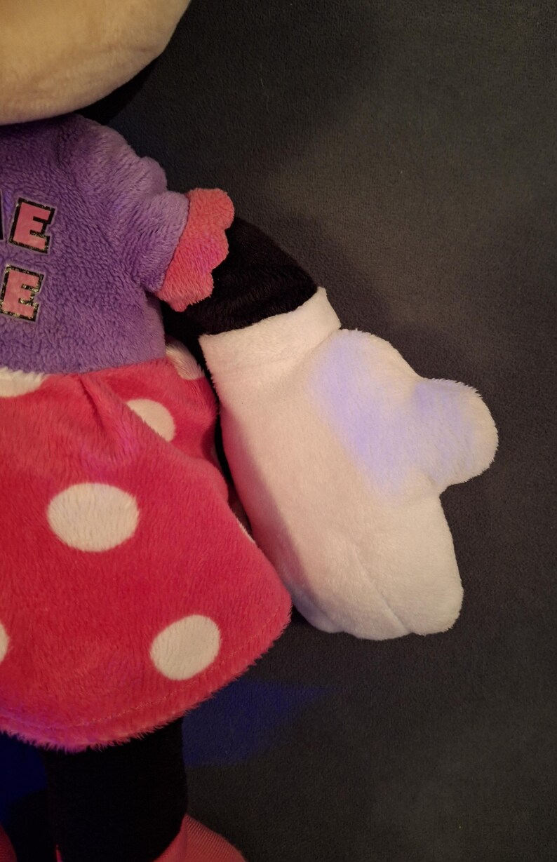 May include: A plush doll featuring a red polka-dot skirt, a purple top with the letter "E" on it, and a white glove. The doll's arm is black and white, with a pink ruffled sleeve. The doll is a popular character.