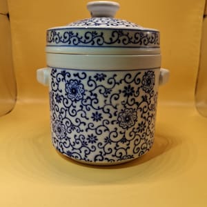 May include: A white and blue ceramic lidded container with a floral pattern. The container has two small handles and a knob on the lid. The floral design is in a dark blue color against a white background. The container is likely for food storage.
