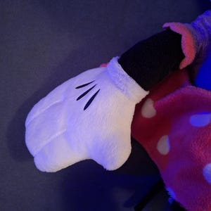 May include: A plush toy featuring a white glove with three black lines on the palm. The glove is part of a larger stuffed character, with a black sleeve and a pink and white polka-dotted garment visible.