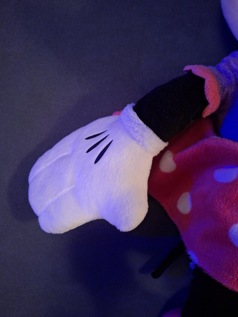 May include: A plush toy featuring a white glove with three black lines on the palm. The glove is part of a larger stuffed character, with a black sleeve and a pink and white polka-dotted garment visible.