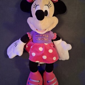 May include: A plush Minnie Mouse doll. The doll has a pink bow with white polka dots, black ears, and a cream-colored face. She wears a purple shirt with "Minnie Mouse" printed on it and a pink skirt with white polka dots. Her shoes are pink.