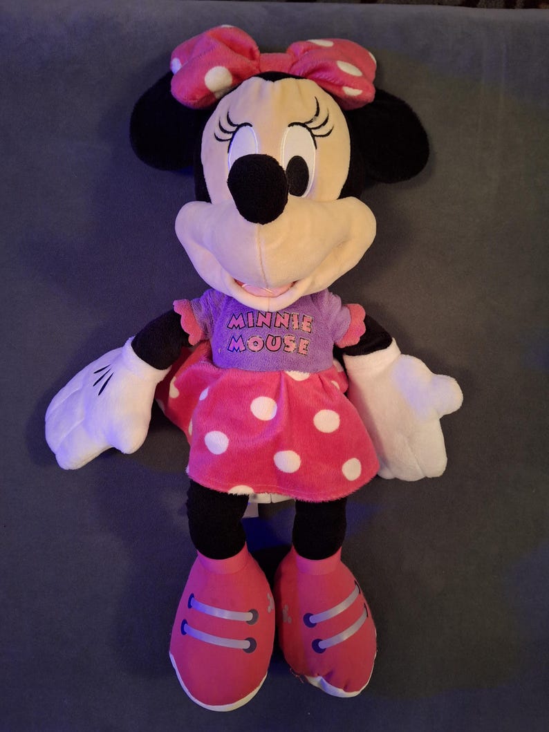 May include: A plush Minnie Mouse doll. The doll has a pink bow with white polka dots, black ears, and a cream-colored face. She wears a purple shirt with "Minnie Mouse" printed on it and a pink skirt with white polka dots. Her shoes are pink.