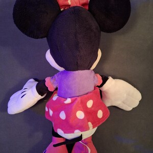 May include: A plush Minnie Mouse doll, viewed from the back, features large black ears, a red polka-dot bow, and a pink polka-dot skirt. The doll has white gloves and pink shoes. The doll is a classic Disney character.