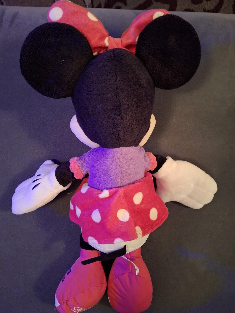 May include: A plush Minnie Mouse doll, viewed from the back, features large black ears, a red polka-dot bow, and a pink polka-dot skirt. The doll has white gloves and pink shoes. The doll is a classic Disney character.
