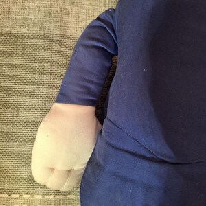 May include: A plush doll with a navy blue suit and white gloves. The doll's arm is visible, showing the sleeve of the suit and the white glove. The background is a textured, light gray surface.