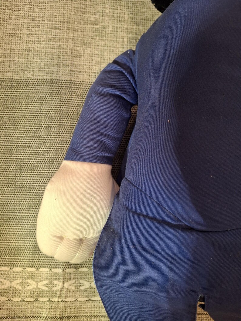 May include: A plush doll with a navy blue suit and white gloves. The doll's arm is visible, showing the sleeve of the suit and the white glove. The background is a textured, light gray surface.