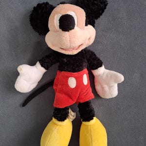 May include: A plush Mickey Mouse toy. The toy features a black body, ears, and tail, with a tan face and white gloves. Mickey wears red shorts with white buttons and yellow shoes. The toy is soft and cuddly.