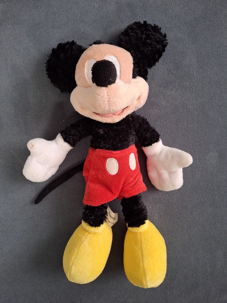 May include: A plush Mickey Mouse toy. The toy features a black body, ears, and tail, with a tan face and white gloves. Mickey wears red shorts with white buttons and yellow shoes. The toy is soft and cuddly.