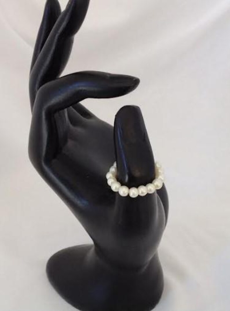 K&ouml;nnte beinhalten: Ein wei&szlig;es Perlenarmband mit schlichtem Design. Das Armband besteht aus kleinen, runden Perlen, die auf einer durchsichtigen Gummikordel aufgereiht sind.