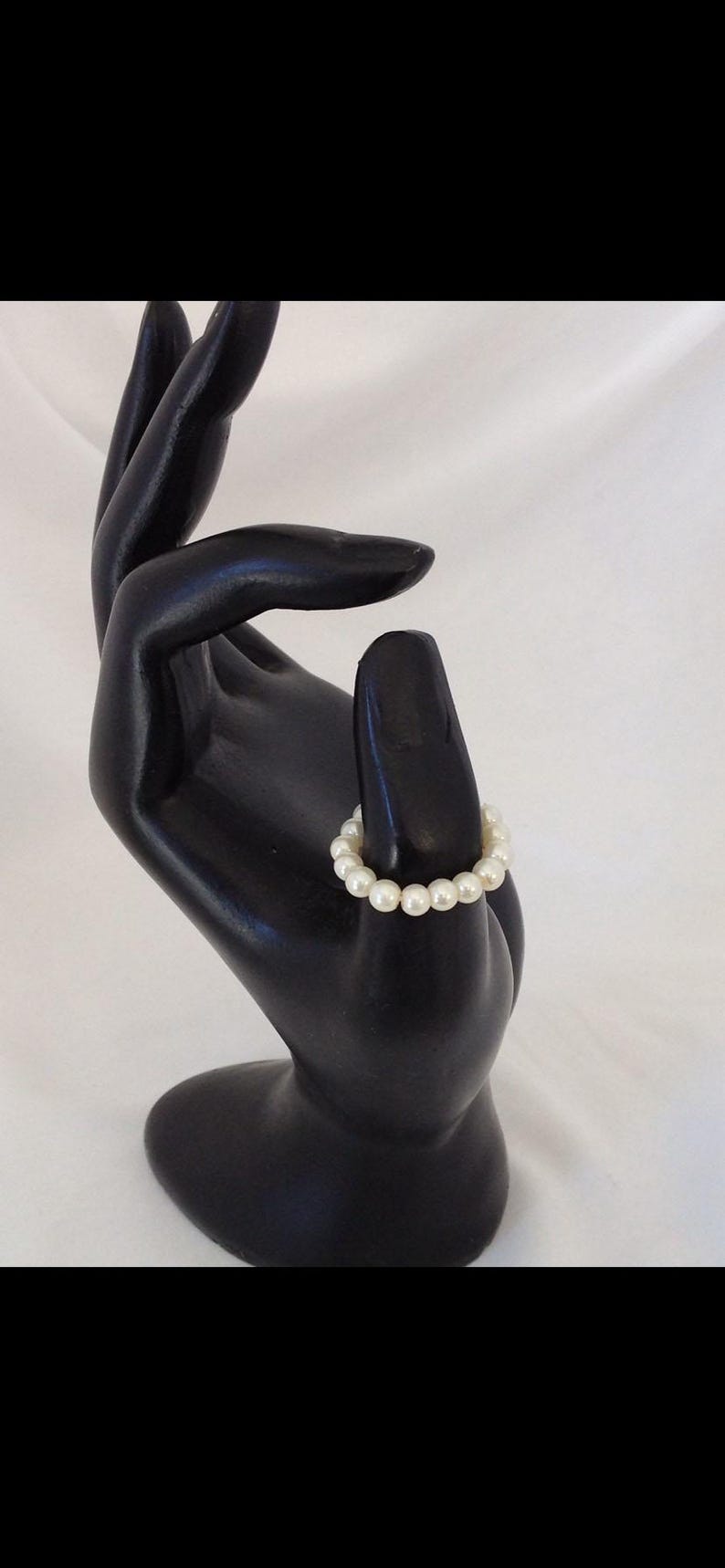 K&ouml;nnte beinhalten: Ein wei&szlig;es Perlenarmband auf einer schwarzen Handpuppe.