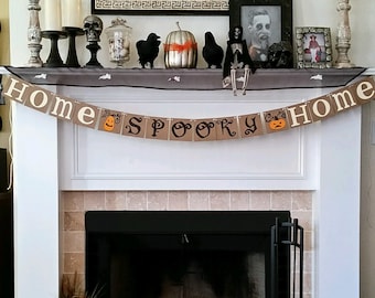 May include: A decorated fireplace mantel with Halloween decorations. A banner reads "HOME SPOOKY HOME" with pumpkin and jack-o-lantern graphics. The mantel displays candles, skulls, crows, a silver pumpkin, and framed pictures. Pumpkins and a lantern are on the hearth.