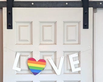 May include: A white banner spelling the word LOVE hangs on a white paneled door. The letter O is replaced by a heart shape featuring horizontal stripes in red, orange, yellow, green, blue, and purple. The door has black metal hardware.