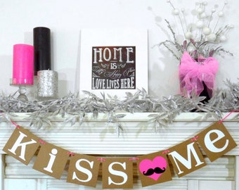 May include: A decorative mantelpiece with a banner that reads "KISS ME". The banner has a pink heart with a black mustache. Other decorations include candles, a framed sign that says "HOME is Happy Place LOVE LIVES HERE", and a vase with flowers.
