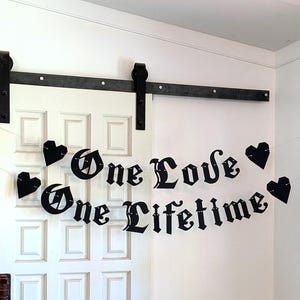 May include: A black hanging banner featuring the text One Love One Lifetime in a gothic-style font. The banner is composed of individual cut-out letters and four black heart-shaped accents, all strung together on a thin, light-colored cord.