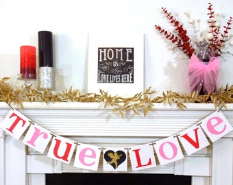 May include: A white mantel decorated for Valentine's Day. A banner reads "True Love" with a heart graphic. Gold garland, candles, and a framed sign that says "Home is where happy is, Love lives here" complete the decor.