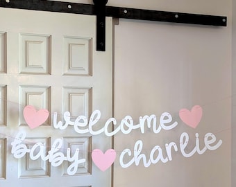 May include: A decorative banner featuring white cursive lettering that spells out "welcome baby charlie" strung on a thin line, accented with three light pink heart-shaped cutouts positioned between the words and at the ends of the text.