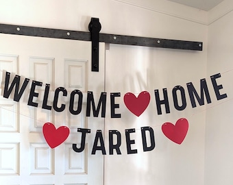 May include: A two-line banner hanging on a string featuring black capital letters that spell out WELCOME HOME JARED. The banner includes three solid red heart-shaped cutouts positioned between the words and at the ends of the second line.