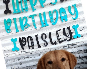 May include: A golden retriever smiles beneath a banner that reads "Happy Birthday Paisley!" The banner features turquoise and black letters, with paw print accents. Bone-shaped cutouts are interspersed throughout the banner, creating a festive atmosphere.