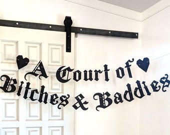 May include: A black paper banner featuring the text A Court of Bitches & Baddies in a gothic-style font, accented with two black heart-shaped cutouts at the ends, all strung together on a thin white cord against a white door background.