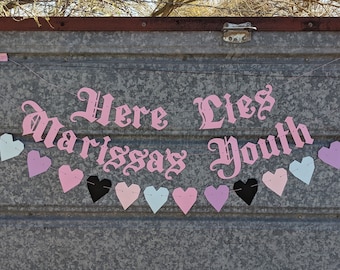 May include: A banner with the text "Here Lies Marissas Youth" in pink gothic lettering, strung with heart-shaped cutouts in black, pink, and light blue. The banner is displayed against a corrugated metal background.