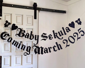 May include: Black banner with the words "Coming March 2025 Baby Sekula" in gothic font, strung across a white door. The banner includes heart and skull and crossbones shapes. Decorative accents hang on the left.