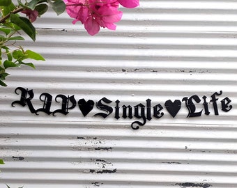 May include: Black metal sign with the phrase "RIP Single Life" in a gothic font, with heart symbols. The sign is mounted on a white, weathered, corrugated metal surface, with pink flowers and green leaves in the background.