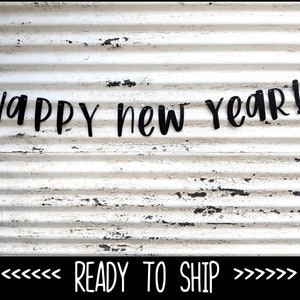 May include: Black "Happy New Year!" banner strung across a weathered, white corrugated metal background. The banner is made of individual black letters. The words "READY TO SHIP" are at the bottom of the image.