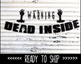 May include: Black Halloween banner with the words "WARNING" and "DEAD INSIDE". The banner includes black zombie hands and a ghost cutout. The banner is displayed against a white, weathered background.