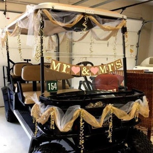 May include: A black golf cart decorated for a wedding with a gold and white tulle and ribbon garland. The cart has a sign that reads 