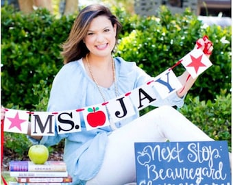 May include: A woman sitting on grass holds a banner that reads "MS. JAY" with a red apple graphic. A sign says "Next Stop: Beauregard Elementary 4th grade." A stack of books and a green apple are also present.