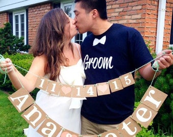 May include: A couple kissing, holding a banner with the names "Ana & Peter" and the date "11-14-15". The banner is made of brown cardboard squares with white lettering and is strung on green twine. The man wears a navy blue shirt with a white bow tie and the word "Groom".