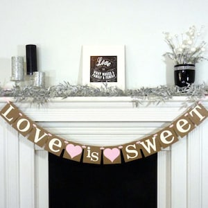 May include: A decorative banner with the words "Love is sweet" in white letters and pink hearts. The banner is strung across a white mantelpiece, with a framed print and floral arrangement in the background.