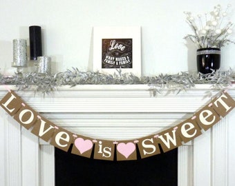 May include: A decorative banner with the words "Love is sweet" in white letters and pink hearts. The banner is strung across a white mantelpiece, with a framed print and floral arrangement in the background.
