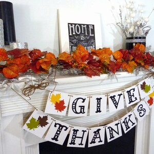 May include: Thanksgiving-themed mantel decoration with a banner that reads "GIVE THANKS." The banner has colorful leaf accents. The mantel also features fall foliage, a framed sign that says "HOME is a happy place LOVE is here," and decorative candles.