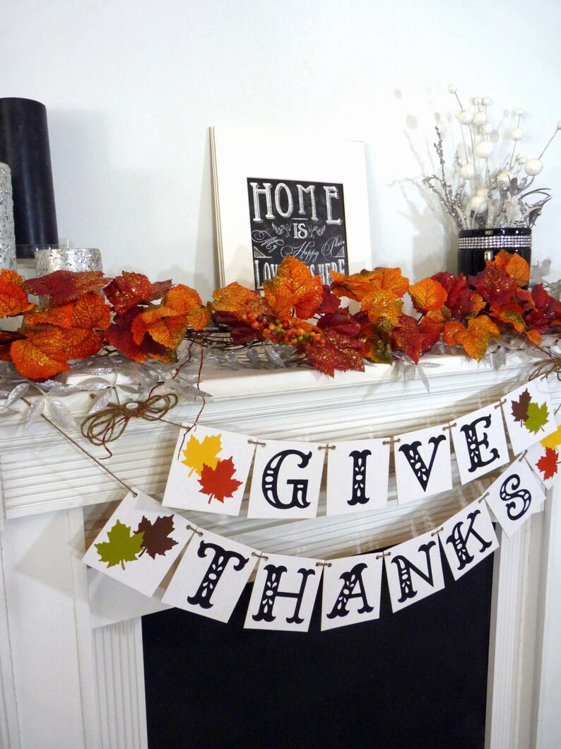 May include: Thanksgiving-themed mantel decoration with a banner that reads "GIVE THANKS." The banner has colorful leaf accents. The mantel also features fall foliage, a framed sign that says "HOME is a happy place LOVE is here," and decorative candles.