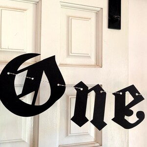 May include: A black paper banner featuring gothic-style lettering that spells out the word One, with a heart-shaped cutout on the left side. The individual letters and the heart are strung together on a thin white string for hanging display.